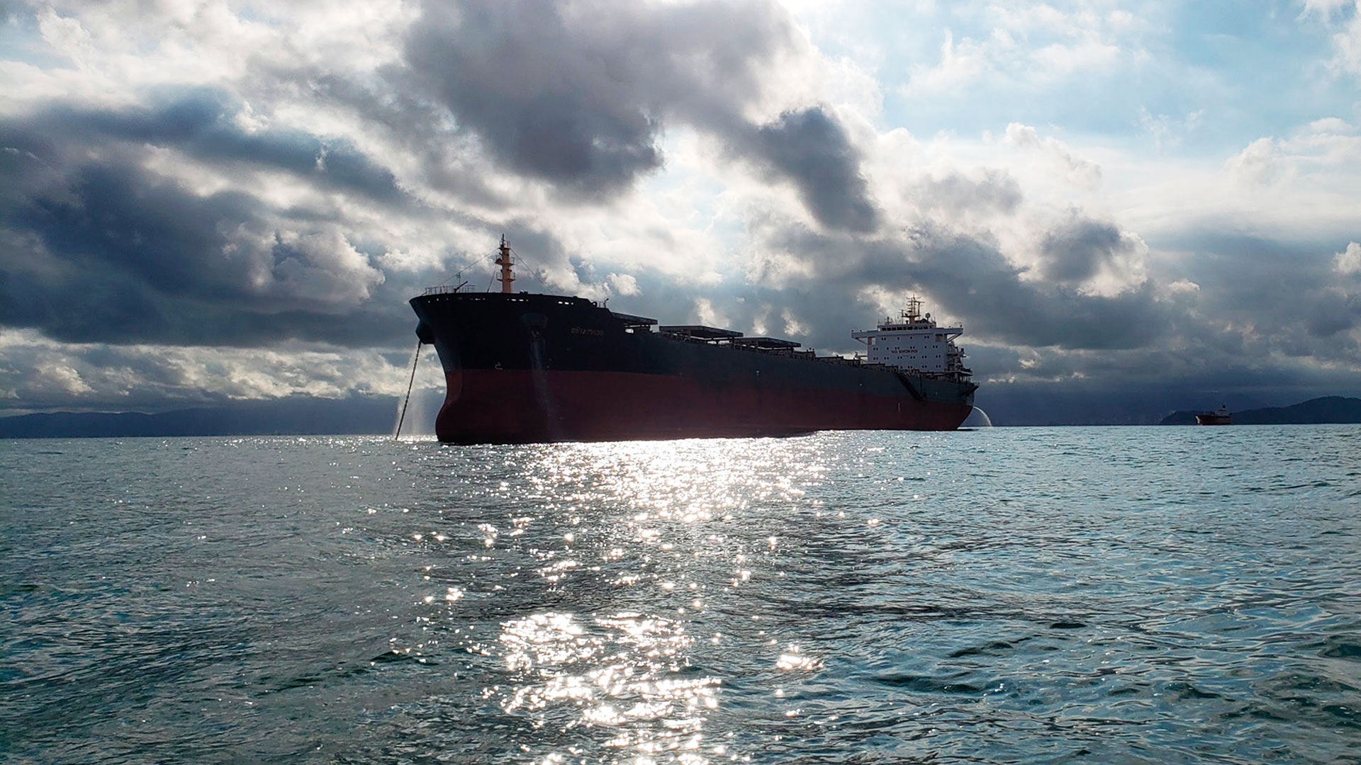 Navio cargueiro fotografado no oceano. O navio é vermelho e preto. O céu está claro com muitas nuvens e ao fundo é possível ver uma porção de terra