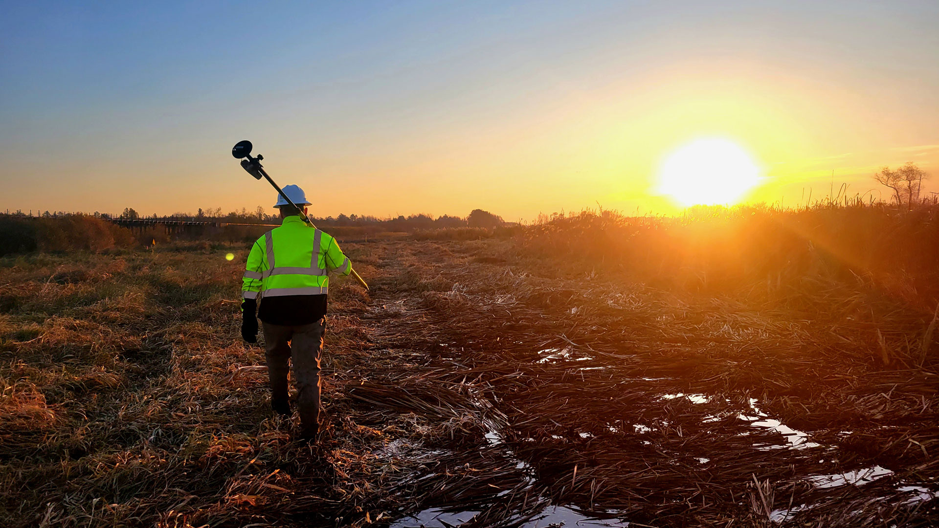 Homem carregando um instrumento de análise ambiental em uma planície com bastante vegetação rasteira. Há um nascer do sol ao fundo