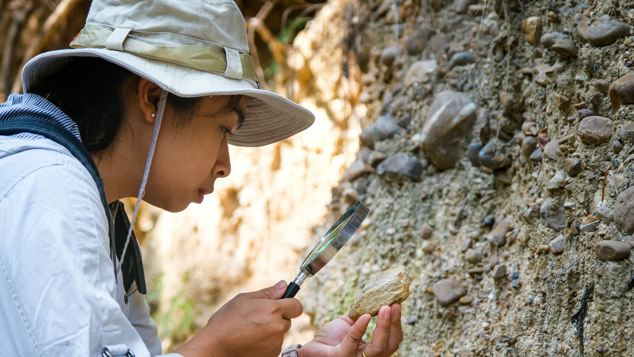 Mulher geologista com chapéu e mochila analisando rocha com uma lupa na mão.