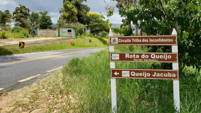 Placa em beira de estrada citando "Circuito Trilha dos Inconfidentes"; "Rota do Queijo" e "Queijo Jacuba".