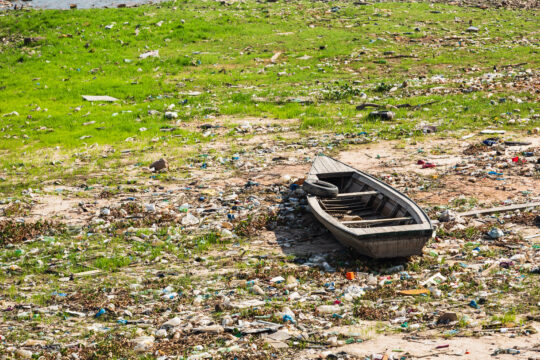 Na região conhecida como Manaus Moderna, em meio à seca severa do Rio Negro, barcos estão encalhados na margem. O cenário é marcado por um solo exposto, coberto de lixo e detritos, evidenciando os impactos ambientais da crise hídrica que afeta a Amazônia. A vegetação verde ao redor contrasta com a poluição, refletindo a fragilidade do ecossistema local diante das mudanças climáticas.